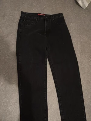 Svarta Supreme jeans straight fit - Supreme jeans i svart denim med klassisk femficksdesign och raka ben. Byxorna har Supreme-detaljer på knapp och röd etikett vid fickan. Tillverkade i 100% bomull för en skön och slitstark känsla. Perfekta för dig som gillar streetwear och vill ha en clean look.