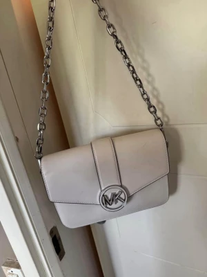 Vit axelväska från Michael Kors - Snygg vit axelväska från Michael Kors med silverfärgad kedjerem och ikoniskt MK-spänne framtill. Väskan har stilren design, raka linjer och är tillverkad i slätt skinn. Invändigt finns ett fack med dragkedja och avtagbar axelrem i matchande färg.