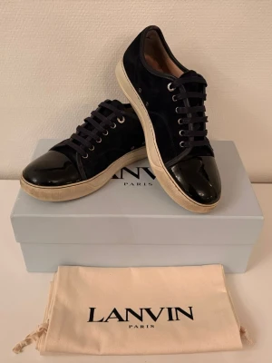 Mörkblåa Lanvins med glans  - Säljer ett par mörkblåa Lanvin sneakers med snygg kontrast mellan mocka och lackad tå. Skorna har vita sulor, svarta snören och klassisk låg profil. Dustbag och originalkartong medföljer.