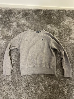 Polo Ralph Lauren grå sweatshirt  - En stilren grå sweatshirt från Polo Ralph Lauren i bomull. Klassisk passform med rund hals, ribbade muddar och det ikoniska blå broderade logotypen på bröstet. Perfekt för en chill och avslappnad look.