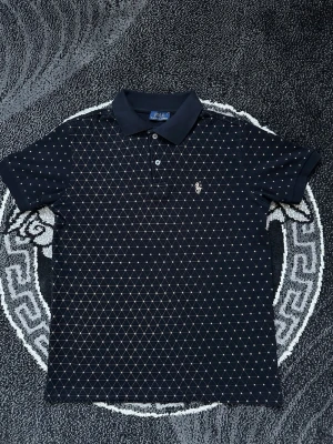 Svart piké från Polo Ralph Lauren - Säljer en svart pikétröja från Polo Ralph Lauren med triangulärt rutmönster i vitt och klassisk krage. Tröjan har korta ärmar, två knappar framtill och den ikoniska broderade loggan på bröstet. Perfekt till en clean och stilren look.