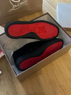 Svarta Louboutin skor - Säljer ett par svarta sneakers från Christian Louboutin med ikonisk röd sula och diskret snörning. Skorna är tillverkade i svart mocka och har en stilren, rund tå. Kommer med originalkartong, dustbags och skohorn.