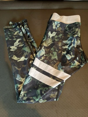 Camo leggings från Stronger, S - Säljer ett par snygga camo-mönstrade leggings från Stronger i storlek S. De har bred vit midjeresår och två vita ränder på ena benet. Materialet är stretchigt och består av 82% polyester och 18% elastan, perfekta för träning eller chill.