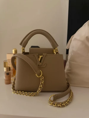 Beige handväska  med gulddetaljer  - En elegant och tidlös handväska i en varm, beige ton som passar lika bra till vardag som till mer uppklädda tillfällen. Väskan är tillverkad i strukturerat läder med en gedigen känsla och fina sömmar som ger ett exklusivt uttryck. Den stabila topphandtaget gör den bekväm att bära i handen, medan den avtagbara och justerbara axelremmen med dekorativ kedjedetalj ger flexibilitet i användningen. Guldfärgade metalldetaljer ger väskan en sofistikerad kontrast och förstärker helhetsintrycket. 