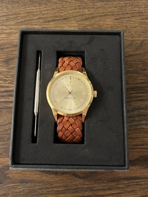Triwa Lansen - En guldig klocka med en guldig urtavla och ett flätat brunt klockarmband. Sprillans ny och helt oanvänd. Storlek 38 urtavla. 