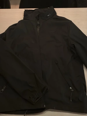 Svart vindjacka från GANT XL - Säljer en svart vindjacka från GANT i storlek XL. Jackan har hög krage, dragkedja framtill och två snedställda fickor med dragkedja. Materialet är slätt och känns sportigt, perfekt för blåsiga dagar. Enkel och stilren design med diskret GANT-logga på bröstet.