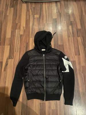 Svart Moncler pufferjacka med huva - Säljer en svart pufferjacka från Moncler med huva och dragkedja framtill. Jackan har stickade ärmar och en stor vit Moncler-logga på ena ärmen. Kroppen är quiltad och har två fickor med knappstängning. Perfekt för dig som vill ha en snygg och sportig look.