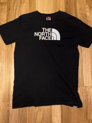 Svart t-shirt från The North Face - Svart t-shirt från The North Face med stor vit logga på bröstet. Klassisk rund halsringning och korta ärmar. Tillverkad i mjuk bomull som är skön att ha på sig. Perfekt för dig som gillar streetwear och enkel stil.