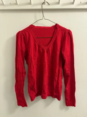 Röd v-ringad tröja med puffärm - Röd långärmad tröja med v-ringning och puff vid axlarna. Ärmsluten har snygga knappar som detalj. Tröjan är i mjuk bomull och har en figurnära passform. Perfekt för dig som vill ha en enkel men ändå trendig look.