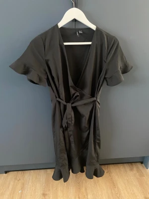 Svart omlottklänning  - Säljer en svart omlottklänning från Vero Moda i storlek S. Klänningen har korta volangärmar, knytband i midjan och volangkant nertill. Materialet är mjukt och lätt, perfekt för en trendig look. Djup v-ringning framtill ger en snygg siluett.