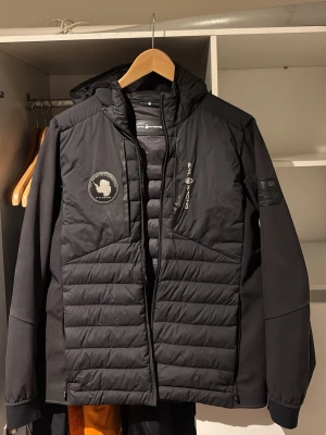 Svart Sail Racing dunjacka med huva - Säljer en svart Sail Racing JR Antarctica Hybrid Hood med huva. Jackan har quiltade partier, dragkedja framtill och logga på bröstet . Perfekt för kyliga dagar och snygg streetstil.