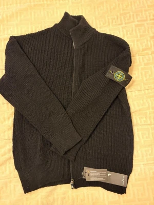 Stone Island stickad zip-tröja – svart – XL – helt ny med tags - Helt ny Stone Island stickad tröja med hel dragkedja och klassiska Stone Island-patchen på vänster arm. Aldrig använd, alla tags sitter kvar. 	•	Storlek: XL 	•	Färg: Svart 	•	Modell: Stickad zip-tröja / cardigan 	•	Skick: Helt ny 	•	Originalpatch med knappar  Perfekt för både vardag och mer uppklädda outfits. Skickas snabbt och säkert. Hör av dig vid frågor eller om du vill ha fler bilder.
