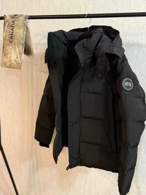 Svart pufferjacka från Canada Goose - Säljer en svart pufferjacka från Canada Goose med avtagbar huva och patch på ärmen. Jackan har dragkedja och knappar framtill, samt medföljande pälsdetalj till huvan. Insidan har praktiska axelband och meshfickor. Perfekt för kalla vinterdagar. Finns i alla storlek