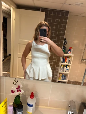 Vit peplum linnetopp med rosett - Säljer en vit peplum linnetopp med breda axelband och öppen rygg. Toppen har en snygg rosett och knytning baktill samt en figurnära passform upptill som blir utsvängd nertill. Perfekt för dig som vill ha en stilren och trendig look. Aldrig använd! Nypris 400kr