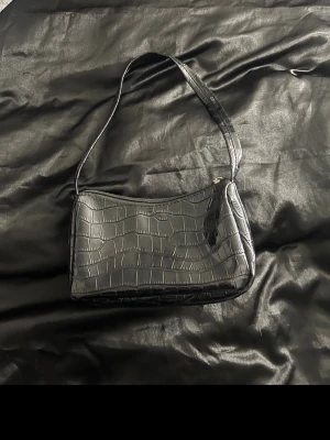 Svart axelväska i krokopräglat skinn - Snygg svart axelväska i krokopräglat skinnimitation med enkel dragkedja upptill. Väskan har en stilren, rektangulär form och ett smalt axelband som gör den enkel att bära. Perfekt accessoar för att lyfta din outfit.