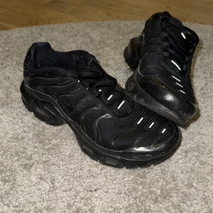 Nike Air Max Plus svarta sneakers - Nike Air Max Plus sneakers i helsvart med mesh och syntetpaneler, ikoniska vågiga linjer och synlig Air-enhet i sulan med gula detaljer. Skorna har snörning och reflekterande inslag på ovansidan. Perfekt för dig som gillar streetwear och sportig stil.