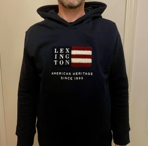Mörkblå  Lexington hoodie med broderi - Mörkblå hoodie från Lexington med broderad patch framtill i rött, vitt och blått. Snyggt tryck med texten 'American Heritage Since 1993'. Tröjan har huva och långa ärmar, tillverkad i mjuk bomull. Perfekt för dig som gillar klassisk och stilren streetwear.Helt ny me prislapp,original pris 895 mitt pris 599kr.De är storlek 170 men den passar S.