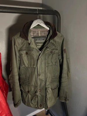 Grön parkajacka från Marc O'Polo - Snygg grön parkajacka från Marc O'Polo med quiltat foder och flera praktiska fickor framtill. Jackan har en mörkbrun krage i manchester, dragkedja och knappar. Perfekt för dig som gillar en klassisk och chill stil med funktionella detaljer.