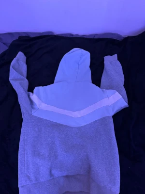 Grå och ljusblå hoodie med emblem - Säljer en snygg trapstar hoodie i grått och ljusblått med vita detaljer. Tröjan har en huva med snörning, ett broderat emblem på bröstet och en stor magficka. Perfekt för chill dagar och streetwear-stil.