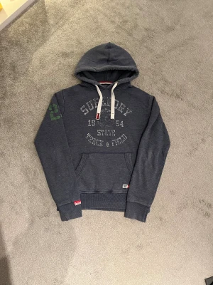 Mörkblå Superdry hoodie - Riktigt fet mörkblå hoodie från Superdry med stort tryck på bröstet och grönt 23-tryck på ena ärmen. Tröjan är i storlek M och  har riktigt bra passform. 
