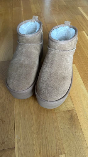 UGG Ultra Mini Platform - UGG boots i chestnut med fluffigt vitt foder och rejäl platåsula i storlek 41. Skorna har rund tå och låg modell, perfekta för kalla dagar. Klassisk UGG-logga bak och dragflik för enkel påtagning. Snyggt och mysigt val för höst och vinter. Inköpta i december förra året. Sparsamt använda men har lyckats få några skråmor i plattformen som finns att se på bilderna, men inget som påverkar mer än det kosmetiska. Skickas i originalkatongen. Kan gå ner i pris vid snabb affär