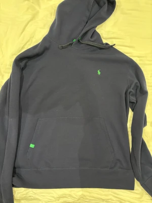 Mörkblå XL hoodie från Polo Ralph Lauren - Mörkblå hoodie från Polo Ralph Lauren med klassisk grön logga på bröstet. Tröjan har huva med snörning, stor magficka och är tillverkad i mjuk bomull. Perfekt för chill dagar och enkel att matcha med jeans eller joggers. Storlek XL.