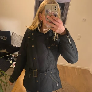 Svart quiltad jacka från Barbour - Quiltad svart jacka från Barbour International med bälte i midjan, dragkedjor och knappar i guldton. Jackan har hög krage med mysig foderkant och flera fickor framtill. Perfekt för dig som vill ha en klassisk men edgy look.