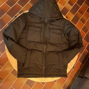 Svart pufferjacka från Jack & Jones - Svart pufferjacka från Jack & Jones med huva och två stora bröstfickor med lock. Jackan har dragkedja framtill och är quiltad för extra värme. Perfekt för kalla dagar och har en clean, stilren look som funkar till det mesta.