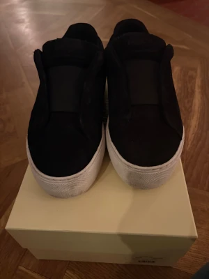 Svarta sneakers från Axel Arigato - Svarta slip-on sneakers från Axel Arigato med vit platt sula och rund tå. Skorna har en stilren design med bred elastisk rem över foten och logga på plösen samt undersidan. Ovandelen är i mocka och sulan i gummi, perfekt för dig som gillar minimalistisk streetstil.