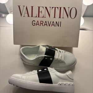 Valentino Garavani vita sneakers - Snygga vita sneakers från Valentino Garavani med bred svart rem över sidan och coola nitar på hälen. Skorna har snörning, platt sula och är tillverkade i lyxigt skinn. Perfekt för dig som gillar stilrena och exklusiva detaljer.