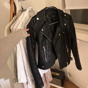 Svart skinnjacka från Vero Moda - Säljer en svart skinnjacka från Vero Moda med silvriga dragkedjor och knappar. Jackan har klassisk bikerstil med bred krage och detaljerade sömmar på ärmarna. 