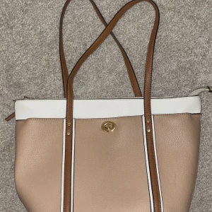 Beige och vit axelväska  - Snygg axelväska i beige och vitt skinn med bruna axelremmar. Väskan har guldfärgad vridknapp och dragkedja upptill. Stilren design med kontrasterande färgpaneler och detaljer i guld. Perfekt storlek för att få plats med allt du behöver.
