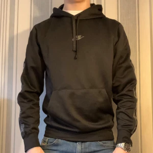 Svart Nike hoodie med loggan i mitten - Svart hoodie från Nike med loggan i mitten och skönt material. Tröjan passar perfekt nu till vintern och har även en snygg luva. Tröjan har Nike text på armarna. Tröjan är äkta och väldigt bra skick!