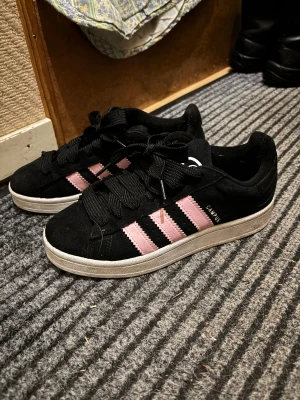 Adidas Campus sneakers svart/rosa - Säljer ett par Adidas Campus sneakers i svart mocka med klassiska rosa ränder på sidorna och vit sula. Skorna har rund tå, breda svarta skosnören och Adidas logga på tungan och hälen. Perfekta för dig som vill ha en stilren men ändå färgglad sneaker.