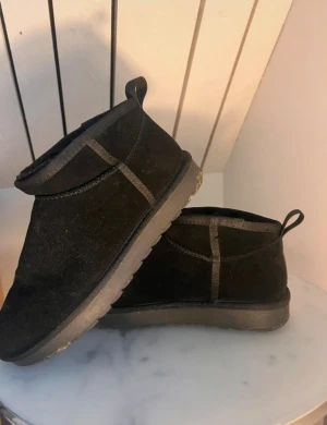 Svarta mockaboots med platt sula - Svarta boots i mocka med platt sula och rund tå. Skorna har en låg siluett och dragflik baktill för enkel påtagning. Yttersulan är mönstrad för extra grepp, perfekt för höst och vinter. Stilrena och passar till många olika outfits.