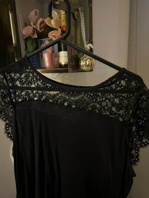 Svart klänning med spetsdetaljer - Svart tröja med snygga spetsdetaljer över axlar och bröst. Modellen har rund halsringning och korta ärmar med spets som ger en cool vibe. Materialet känns mjukt och faller fint, perfekt för dig som gillar stilrena plagg med lite edge.