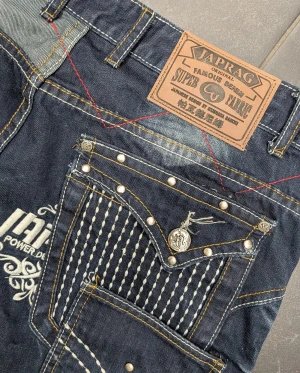 Japrag JP-111D jeans med tryck och nitar - Unika mörkblå jeans från Japrag JP-111D med vita tryck, kontrastsömmar, nitar och flera dragkedjor. Snygga detaljer på fickorna, broderade mönster och coola texttryck. Passar dig som vill sticka ut med en streetwear-look. Materialet är klassisk denim.  Mått: Midjemått: 43 cm (mätt platt) Höft: ca 56 cm Innerbenslängd: 84 cm Bredd vid låret: 30 cm Skrev: 24 cm Stuss: 37 cm Ytterbenslängd: 113 cm Benvidd längst ner: 20 cm  Skriv för frågor, pris kan diskuteras ❤️  