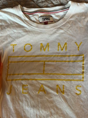 Vit t-shirt Tommy Jeans XS - Vit t-shirt från Tommy Jeans i storlek XS med gul tryckt logga på bröstet och klassisk rund hals. T-shirten har korta ärmar och är gjord i mjuk bomull. Liten broderad Tommy Jeans-flagga på ena ärmen för extra detalj.