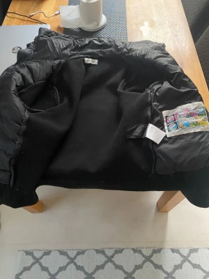 Svart Moncler pufferjacka med ficka - Svart pufferjacka från Moncler med stickade partier och quiltad front. Jackan har en praktisk ficka med dragkedja på ärmen och Moncler-logga. Insidan har en cool serietidningsetikett och QR-kod för äkthetskontroll. Perfekt för dig som gillar exklusiva streetwear-vibbar.