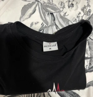 Svart Moncler t-shirt med logga - Svart t-shirt från Moncler i 100% bomull med klassisk rund hals och korta ärmar. Stor Moncler-logga i vitt och rött framtill ger en clean och stilren vibe. Perfekt för dig som gillar exklusiva märken och enkel design.