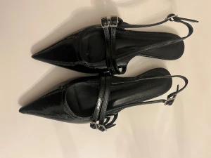 Svarta spetsiga skor med bälten - Snygga svarta finskor med spetsig tå och dubbla tunna remmar med silverfärgade spännen. Skorna har öppen häl och platt sula, tillverkade i glansigt material. Perfekta för dig som gillar en stilren och edgy look.