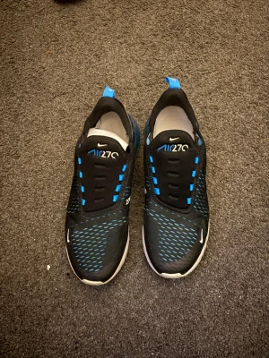 Nike Air 270 sneakers svart/blå - Nike Air 270 sneakers i svart med blå detaljer och vit sula. Skorna har en luftig mesh-överdel, tydlig Air Max-bubbla i hälen och blå accenter på snörningen och loggan. Perfekta för dig som gillar sportig och modern stil.