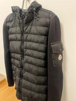 Svart Moncler pufferjacka med huva - Svart pufferjacka från Moncler med stickade ärmar och quiltad kropp. Jackan har huva med snörning, dragkedja framtill och en ficka med Moncler-logga på ärmen. Perfekt för dig som vill ha en clean och stilren look med premiumkänsla.