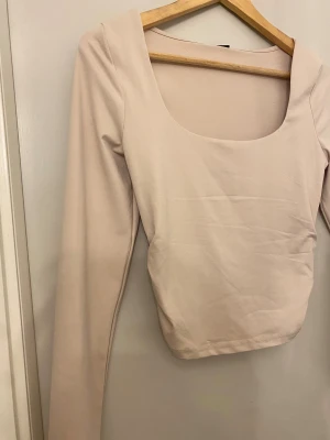 Beige långärmad topp med fyrkantig hals - Snygg beige långärmad topp med en bred fyrkantig halsringning. Modellen är figurnära och har en stilren, minimalistisk look. Perfekt att matcha med jeans eller kjol för en clean vibe. Materialet känns mjukt och stretchigt, vilket gör den skön att bära.