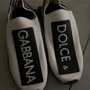 Dolce & Gabbana vita sneakers - Säljer ett par vita Dolce & Gabbana sneakers med svart och vit kontrastdesign. Skorna har en bred svart panel med stor logotyp på ovansidan, elastisk ovandel i syntetmaterial och en chunky svart sula. Snygga detaljer med logoband runt hälen och platt sula för skön komfort.