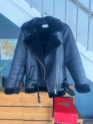 Svart teddyjacka från NA-KD med bälte - Säljer en svart teddyjacka från NA-KD i storlek 34. Jackan har bred krage, mjukt fuskpälsfoder, detaljer med bälte och silverfärgad dragkedja. Perfekt för dig som gillar en cool och mysig look under höst och vinter.