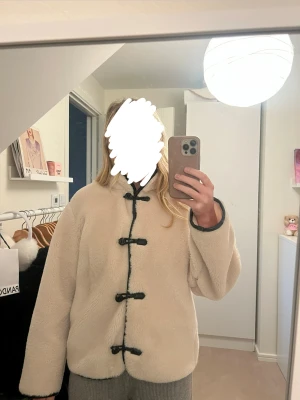 Beigevit teddyjacka med snörning - Säljer en beige teddyjacka med svarta snörningsdetaljer framtill. Jackan har en loose passform och långärmade ärmar. Väldigt skön att ha på sig när det är kallt utomhus.