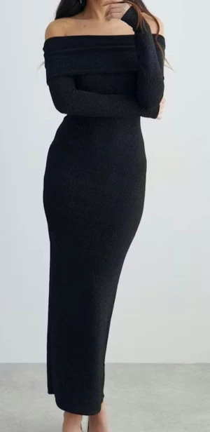 Klänning  - Supersnygg svart långklänning med glittrigt tyg och offshoulder-design. Klänningen har lång ärm, figurnära passform och en slits bak för extra edge. Perfekt för dig som vill sticka ut med en elegant och trendig look. Materialet är glittrigt syntet som ger en lyxig känsla.