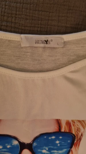 Vit linnetopp med färgglatt tryck - Säljer en vit linnetopp i storlek L med ett stort färgglatt tryck framtill. Trycket har motiv med solglasögon, vattenmelon, badring och drink, vilket ger en somrig vibe. Toppen är ärmlös och har rund hals. Perfekt för dig som gillar statement-plagg.