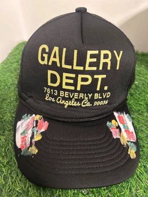 Svart keps Gallery Dept mesh - Svart truckerkeps från Gallery Dept. med mesh baktill och justerbar snapback. Framsidan har gul text och handmålade färgdetaljer i rött, grönt, vitt och gult på skärmen. Snygg och unik accessoar med streetkänsla.